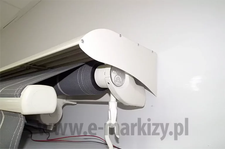 Zawieszona na białej ścianie markiza firmy MOL z białą konstrukcją, otwieraniem ręcznym za pomocą korby zaczepionej do uchwytu oczkowego po prawej stronie markizy, z lekko wysuniętą belką przednią z poszyciem i falbaną wykonaną z tkaniny w kolorze ciemnej szarości z wąskimi paskami w kolorze białym oraz białym obszyciem, nad którą zamontowany jest biały daszek ochronny do markiz MOL, pod markiza widoczne dwa czarno-czerwone przewody do oświetlenia do markizy