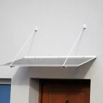 Prosty, klasyczny daszek nad drzwi Sewilla w kolorze białym, zamocowany na odciągach do jasnej elewacji. Pod daszkiem widoczna wnęka z brązowymi drzwiami, a po lewej stronie okno z białą ramą.