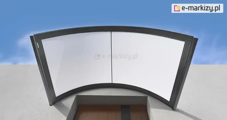 Grafika przedstawia widok z góry na nowoczesny daszek naddrzwiowy o łukowatym kształcie, zamontowany nad wnęką wejściową budynku. Konstrukcja zadaszenia składa się z ciemnoantracytowej ramy aluminiowej, która dzieli powierzchnię na dwa symetryczne panele. Wypełnienie paneli stanowi biały, półtransparentny poliwęglan komorowy, który skutecznie rozprasza światło. Rama daszka posiada wyraźnie zaznaczone profile krawędziowe oraz pionowy szpros przebiegający dokładnie przez środek konstrukcji. Daszek jest zamontowany do jasnej, płaskiej elewacji i wystaje poza obrys wnęki drzwiowej, widocznej w dolnej części kadru. Wnęka ta ukazuje górny fragment drzwi o brązowym kolorze z pionowym, ciemniejszym pasem dekoracyjnym. Całość prezentowana jest na tle czystego, błękitnego nieba z delikatnymi smugami białych chmur po bokach. Perspektywa od dołu pozwala dokładnie zaobserwować łukowate wygięcie daszka oraz sposób, w jaki jego krawędzie są wykończone profilami odprowadzającymi wodę opadową na boki.