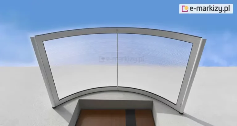 Widoczny od dołu zamocowany nad drzwiami wejściowymi niskiego budynku daszek nad drzwi o łukowym kształcie z aluminiową konstrukcją w kolorze białym oraz wypełnieniem z przejrzystego poliwęglanu komorowego. Nad budynkiem i przez płytę daszka widoczne niebieskie niebo.