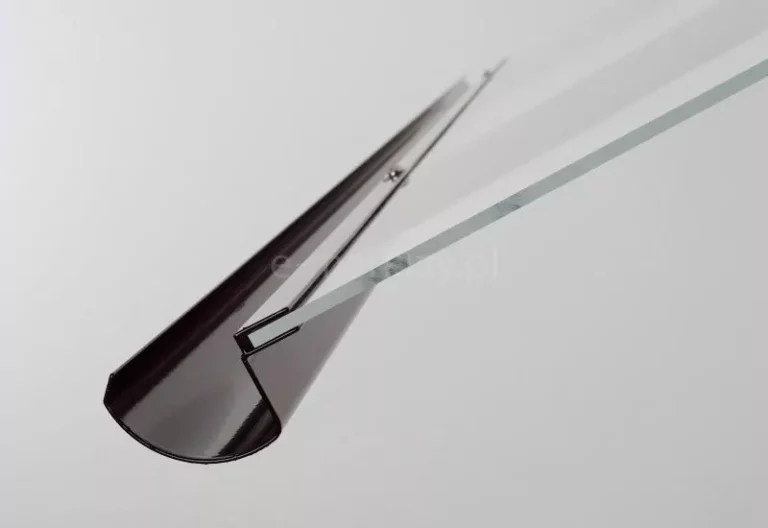 Zbliżenie na detal konstrukcyjny daszka akrylowego Imperia nad drzwiami wejściowymi ukazujące sposób montażu tafli akrylu w profilu aluminiowym o zaokrąglonym, rynnowym kształcie. Profil wykończony jest w ciemnym, brązowym kolorze o wysokim połysku, a w jego wewnętrznym wyżłobieniu osadzona jest krawędź przezroczystej tafli pokrycia daszka o wyraźnie zaznaczonej grubości. Zdjęcie wykonane z perspektywy ukośnej podkreśla precyzyjne dopasowanie elementów mocujących oraz widoczny punkt kotwiczenia śruby montażowej wewnątrz profilu, co świadczy o stabilności i estetyce systemu zadaszenia szklanego. Jasne tło pozwala na dokładną obserwację profilu aluminiowego oraz krawędzi akrylu, eksponując techniczne aspekty instalacji daszka Imperia w warunkach studyjnych.