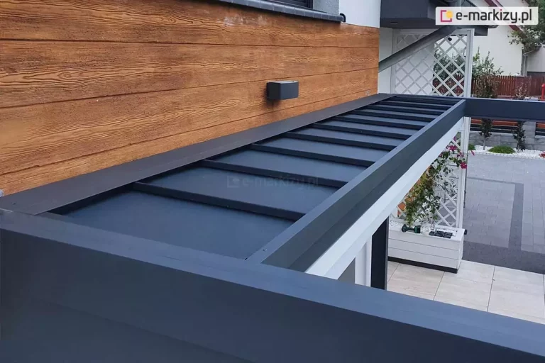 Zdjęcie prezentuje z bliska górny detal konstrukcyjny aluminiowej pergoli tarasowej Luxury w kolorze antracytowym, skupiając się na systemowym daszku ochronnym. Daszek ten jest zamontowany wzdłuż ściany budynku i składa się z kilku płaskich paneli z lekkim spadkiem, które mają za zadanie chronić zwinięty materiał zadaszenia lub mechanizm przed opadami atmosferycznymi i zabrudzeniami. Konstrukcja przylega do elewacji wykończonej dekoracyjnymi deskami drewnopodobnymi w ciepłym, miodowym odcieniu, na której zamontowano nowoczesny, prostokątny kinkiet zewnętrzny w kolorze szarym. W tle, pod konstrukcją pergoli, widoczna jest jasna podłoga tarasowa, biała donica ogrodowa oraz dekoracyjna biała kratka (trejaż) przy ścianie. W oddali widać fragment podjazdu z kostki brukowej oraz ogrodzenie posesji. Kadr zdjęcia podkreśla precyzję łączeń aluminiowych profili oraz estetykę wykończenia zabudowy tarasu.