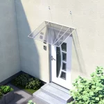 Płaski daszek nad drzwi wejściowe z białą aluminiową konstrukcją zamocowaną do lekko żółtej ściany na odciągach. Grafitowe wypełnienie z poliwęglanu zacienia białe drzwi z ozdobnymi szybkami. Przed wejściem widoczne dwa betonowe stopnie i niewielki ogródek z funkiami i roślinami w donicy.
