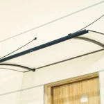 Daszek markizowy nad drzwi o charakterystycznym wygiętym w przód kształcie, z antracytową konstrukcją i przejrzystym wypełnieniem z poliwęglanu, zamocowany do jasnożółtej ściany nad drewnianymi drzwiami za pomocą odciągów.