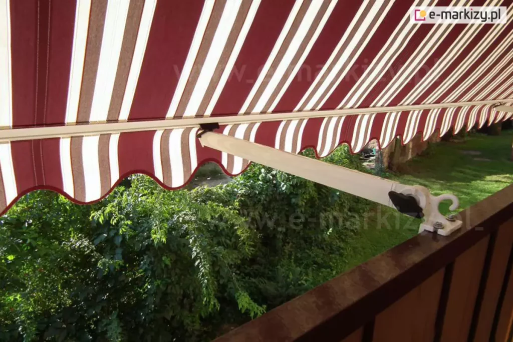 Zbliżenie na markizę balkonową z ramionami poręczowymi, zamontowaną na poręczy brązowej balustrady balkonu. Poszycie markizy wykonane jest z materiału w klasyczne, pionowe pasy w kolorach bordowym, białym i beżowym, zakończone u dołu ozdobną, falowaną falbaną z białą lamówką. Konstrukcja wspiera się na jasnym, kremowym ramieniu, które jest połączone z balustradą za pomocą metalowego uchwytu z pokrętłem regulacyjnym i hakiem. Widok z perspektywy balkonu ukazuje gęstą, soczystą zieleń drzew i krzewów znajdujących się w ogrodzie poniżej, co tworzy naturalne tło dla kolorowego materiału. Światło słoneczne przenika przez tkaninę, podkreślając jej teksturę oraz tworząc wyraźną strefę cienia na balkonie. Kadr skupia się na detalu mechanizmu ramienia oraz estetyce tradycyjnego wzornictwa osłony przeciwsłonecznej.