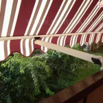 Zbliżenie na markizę balkonową z ramionami poręczowymi, zamontowaną na poręczy brązowej balustrady balkonu. Poszycie markizy wykonane jest z materiału w klasyczne, pionowe pasy w kolorach bordowym, białym i beżowym, zakończone u dołu ozdobną, falowaną falbaną z białą lamówką. Konstrukcja wspiera się na jasnym, kremowym ramieniu, które jest połączone z balustradą za pomocą metalowego uchwytu z pokrętłem regulacyjnym i hakiem. Widok z perspektywy balkonu ukazuje gęstą, soczystą zieleń drzew i krzewów znajdujących się w ogrodzie poniżej, co tworzy naturalne tło dla kolorowego materiału. Światło słoneczne przenika przez tkaninę, podkreślając jej teksturę oraz tworząc wyraźną strefę cienia na balkonie. Kadr skupia się na detalu mechanizmu ramienia oraz estetyce tradycyjnego wzornictwa osłony przeciwsłonecznej.