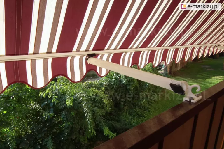 Zbliżenie na markizę balkonową z ramionami poręczowymi, zamontowaną na poręczy brązowej balustrady balkonu. Poszycie markizy wykonane jest z materiału w klasyczne, pionowe pasy w kolorach bordowym, białym i beżowym, zakończone u dołu ozdobną, falowaną falbaną z białą lamówką. Konstrukcja wspiera się na jasnym, kremowym ramieniu, które jest połączone z balustradą za pomocą metalowego uchwytu z pokrętłem regulacyjnym i hakiem. Widok z perspektywy balkonu ukazuje gęstą, soczystą zieleń drzew i krzewów znajdujących się w ogrodzie poniżej, co tworzy naturalne tło dla kolorowego materiału. Światło słoneczne przenika przez tkaninę, podkreślając jej teksturę oraz tworząc wyraźną strefę cienia na balkonie. Kadr skupia się na detalu mechanizmu ramienia oraz estetyce tradycyjnego wzornictwa osłony przeciwsłonecznej.