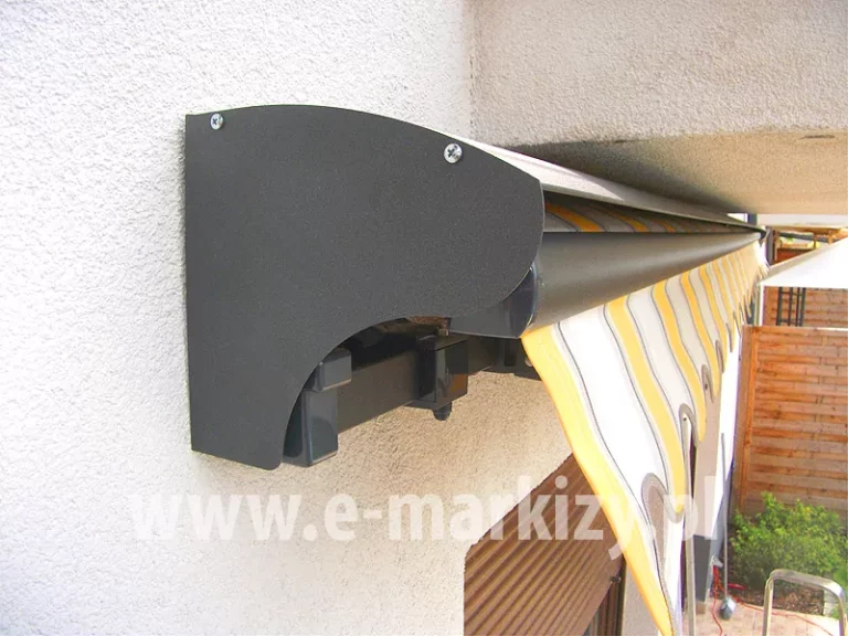Boczek daszka do markiz 22,5cm w kolorze srebrzysty grafit zamocowanego na ścianie z białym tynkiem strukturalnym, osłania rurę nawojową z tkaniną markizy o poszyciu w pasy białe, żółte i szare. Pod daszkiem widoczna falowana falbana, w tle drewniany płot panelowy, a pod markizą widoczne okno osłonięte roletą zewnętrzną w kolorze brązowym