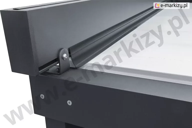 Zbliżenie na przedni narożnik i górną krawędź pergoli Solid Aluprof, prezentujące system uszczelnienia bocznego. Na zdjęciu widoczna jest czarna, gumowa uszczelka o charakterystycznym, piórowym profilu, która jest zamocowana wzdłuż bocznej rynny i szczelnie przylega do jasnej tkaniny zadaszenia, chroniąc wnętrze przed zaciekami wody. Po lewej stronie kadru znajduje się zakończenie profilu bocznego pergoli wykonanego z aluminium w kolorze antracytowym, który jest przytwierdzony do ramy głównej za pomocą solidnego, stalowego wspornika w kształcie litery L, zabezpieczonego śrubą imbusową. Konstrukcja nośna pergoli posiada matowe, strukturalne wykończenie lakiernicze, a na bocznej płaszczyźnie belki widoczne są dwa precyzyjnie osadzone punkty montażowe. Ujęcie to podkreśla techniczne dopracowanie produktu oraz wysoką jakość zastosowanych komponentów uszczelniających, które gwarantują trwałość i funkcjonalność systemu w trudnych warunkach atmosferycznych.