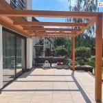 Pergola drewniana Juko przyścienna widok od boku na tarasie pozbawionym mebli i ozdób. W tle widoczny zadbany ogród z trawnikiem i licznymi nasadzeniami niskich drzewek.