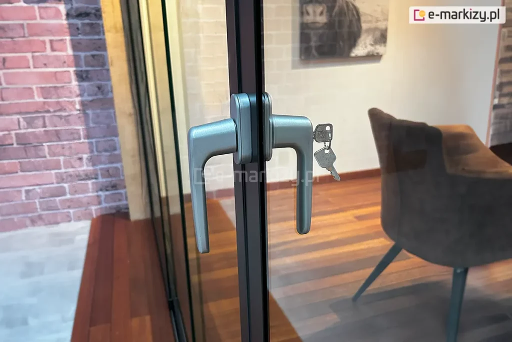 Zbliżenie techniczne na systemowy zamek z klamką dwustronną, zamontowany na profilu krawędziowym szklanych drzwi przesuwnych Auris. Zdjęcie prezentuje ergonomiczną klamkę w kolorze srebrnym/satynowym zintegrowaną z wkładką bębenkową. W zamku umieszczony jest klucz, a drugi kluczyk zwisa na kółku, co podkreśla funkcję zabezpieczenia mienia i możliwość blokowania zabudowy tarasu. Mechanizm jest osadzony na pionowym profilu aluminiowym w kolorze antracytowym, który stabilizuje taflę szkła hartowanego. W rozmazanym tle widoczne jest nowoczesne wnętrze z drewnianą podłogą, stylowym fotelem oraz ścianą z czerwonej cegły.