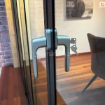 Zbliżenie techniczne na systemowy zamek z klamką dwustronną, zamontowany na profilu krawędziowym szklanych drzwi przesuwnych Auris. Zdjęcie prezentuje ergonomiczną klamkę w kolorze srebrnym/satynowym zintegrowaną z wkładką bębenkową. W zamku umieszczony jest klucz, a drugi kluczyk zwisa na kółku, co podkreśla funkcję zabezpieczenia mienia i możliwość blokowania zabudowy tarasu. Mechanizm jest osadzony na pionowym profilu aluminiowym w kolorze antracytowym, który stabilizuje taflę szkła hartowanego. W rozmazanym tle widoczne jest nowoczesne wnętrze z drewnianą podłogą, stylowym fotelem oraz ścianą z czerwonej cegły.