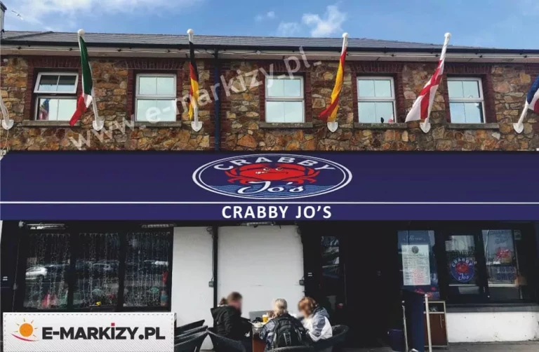 Fotografia przedstawiająca rozłożoną, dużą markizę tarasową zamontowaną nad wejściem do restauracji, ukazana w ujęciu od przodu. Poszycie markizy wykonane jest z gładkiej tkaniny w kolorze ciemnego granatu, tworząc szeroki daszek ochronny nad ogródkiem letnim, w którym przy stolikach siedzą goście. W centralnej części głównej płaszczyzny materiału nadrukowano duże, owalne logo restauracji z podwójną białą obwódką, zawierające rysunkową postać czerwonego kraba, biały napis Crabby u góry oraz pisany odręczną czcionką napis Jo's posadowiony na stylizowanych, niebieskich falach. Przednia krawędź osłony wykończona jest prostą, poziomą falbaną, na której wyśrodkowano duży, czytelny, biały napis Crabby Jo's wykonany prostą czcionką bezszeryfową, podkreślony dodatkowo u góry cienką, poziomą białą linią biegnącą wzdłuż rantu. Powyżej markizy widoczna jest elewacja piętra budynku wykonana z naturalnego, łupanego kamienia w odcieniach brązu i beżu, z pięcioma regularnie rozmieszczonymi oknami w białych ramy. Nad każdym oknem, na białych ukośnych masztach, zamocowane są flagi państwowe różnych krajów, w tym flagi Włoch, Niemiec, Hiszpanii oraz Anglii, swobodnie opadające na tle kamiennej ściany. Pod markizą znajduje się przeszklony front lokalu z ciemnymi ramami okiennymi i wejściowymi, gdzie po prawej stronie widoczne jest menu oraz grafika z logo restauracji naklejona na szybie.