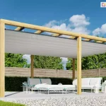 Duża pergola drewniana Juko wolnostojąca ustawiona w ogrodzie w pobliżu drewnianego ogrodzenia oraz niskiego żywopłotu. Konstrukcja na 6 podporach wyposażona w opcjonalny dach rzymski osłania przed słońcem białe meble ogrodowe ustawione na płycie tarasu.