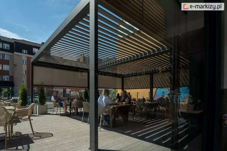 Duża pergola restauracyjna złożona z kilku modułów pergoli Sb400 Aluprof, o konstrukcji aluminiowej w kolorze antracytowym, zainstalowana na letnim tarasie lokalu gastronomicznego. Zadaszenie bioklimatyczne z lamelami ustawionymi pod kątem rzuca charakterystyczny, liniowy cień na podłoże z jasnej kostki brukowej. Ściany boczne pergoli zostały doposażone w rolowane przesłony tkaninowe (refleksole) w kolorze beżowym, które częściowo ograniczają nasłonecznienie. Pod konstrukcją znajdują się klienci siedzący przy stolikach na nowoczesnych, ażurowych krzesłach tarasowych. W tle widoczna jest miejska zabudowa wielorodzinna oraz ulica. Zdjęcie prezentuje zastosowanie profesjonalnych systemów zadaszeń w sektorze komercyjnym (HoReCa) w słoneczny dzień.
