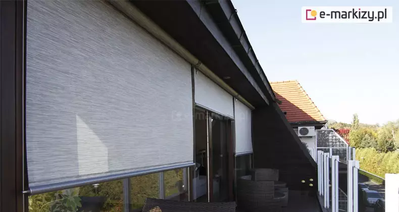 Szerokokątne zdjęcie przedstawiające nowoczesne refleksole XXL Selt zamontowane na przestronnym tarasie lub balkonie apartamentu. Trzy niezależne przesłony w kolorze jasnoszarym o melanżowej strukturze tkaniny są częściowo opuszczone, chroniąc wnętrze przed słońcem i nagrzewaniem. System zamontowany jest pod ciemną podbitką dachową, a jego konstrukcja (prowadnice boczne oraz listwy dolne) wykończona jest w kontrastującym, ciemnym kolorze antracytowym. W tle widoczna jest balustrada tarasowa ze szklanymi panelami, meble ogrodowe z technorattanu oraz panorama osiedla z zielenią i dachami sąsiednich budynków. Zdjęcie prezentuje estetyczny efekt wizualny oraz praktyczne zastosowanie osłon przeciwsłonecznych w architekturze nowoczesnej.