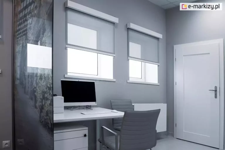 Niewielkie pomieszczenie z ustawionym w narożniku biurkiem z komputerem i białą szafką ukrytą pod blatem oraz dwoma oknami w ścianie za biurkiem i białymi drzwiami po ich prawej stronie. Przy biurku ustawiono dwa szare krzesła, a przy tym odwróconym oparciem do ściany z oknami widoczny jest także biały grzejnik. Oba okna o kwadratowym kształcie zostały osłonięte refleksolami w białych kasetach z tkaniną w odcieniuj jasnej szarości, która została rozwinięta do połowy okien i ogranicza ilość światła wpadającego do pomieszczenia.