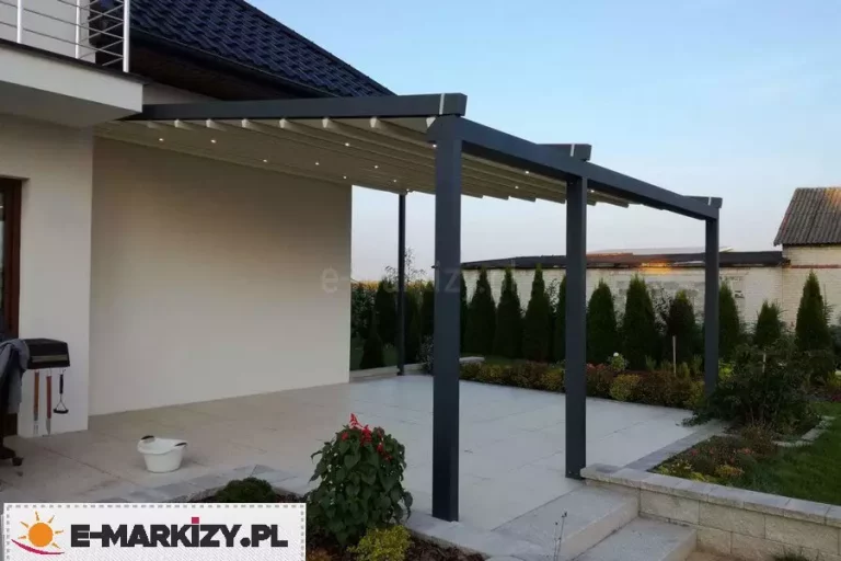 Szeroka, dwumodułowa pergola tarasowa w kolorze ciemnoantracytowym, zamontowana do białej elewacji budynku jednorodzinnego z dachem krytym ciemną dachówką. Konstrukcja wsparta jest na trzech pionowych słupach nośnych i posiada jasne, segmentowe zadaszenie tkaninowe, w którym zintegrowano system oświetlenia LED w postaci punktowych diod rozmieszczonych wzdłuż profili poprzecznych. Pergola osłania przestronny taras wyłożony jasnymi płytami o dużej powierzchni, który od strony ogrodu ograniczony jest niskim murkiem oporowym z betonowych bloczków i rabatami z niską roślinnością. W tle widoczny jest wysoki, gęsty żywopłot z tui oraz fragmenty zabudowy sąsiedniej pod jasnym niebem, co podkreśla nowoczesny charakter aranżacji przestrzeni wypoczynkowej.
