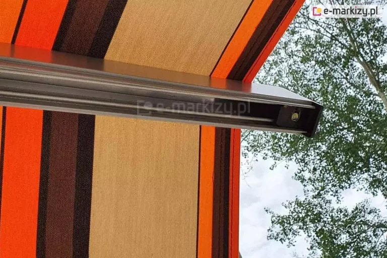 Zbliżenie na techniczny detal konstrukcyjny, jakim jest belka środkowa markizy tarasowej Allegro. Na zdjęciu widoczna jest solidna belka środkowa wykonana z ekstrudowanego aluminium w kolorze srebrnym, która stabilizuje tkaninę i zmienia kąt działania ramion, tworząc charakterystyczne łamanie w połowie jego długości. Materiał markizy charakteryzuje się wyraźnym splotem i wzorem w pionowe pasy o nasyconych barwach pomarańczowego, głębokiego brązu oraz piaskowego beżu. Tkanina jest napięta równomiernie, co świadczy o precyzyjnym działaniu ramion przegubowych. W tle, poza obrysem osłony, widoczne są rozmyte korony drzew liściastych oraz jasne, lekko zachmurzone niebo, co podkreśla funkcję ochronną produktu w warunkach zewnętrznych. Fotografia koncentruje się na wysokiej jakości wykończenia elementów metalowych oraz estetyce tekstury tkaniny akrylowej, odpornej na działanie czynników atmosferycznych.