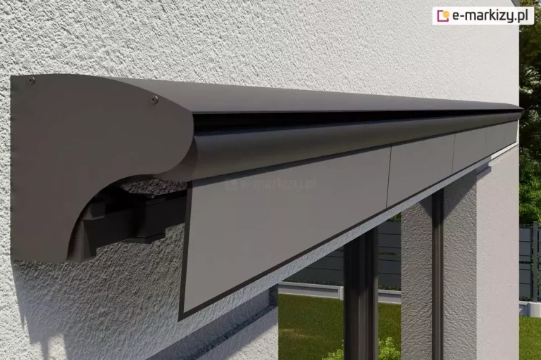 Elegancka grafitowa markiza Jamaica Aluprof z dodatkową osłoną w postaci półkasety/daszka, który chroni tkaninę poszycia. Pod daszkiem widoczna prosta falbana z szarej tkaniny. Pod markizą widoczne duże okno tarasowe, w którym odbija się ogród z zadbanym trawnikiem.