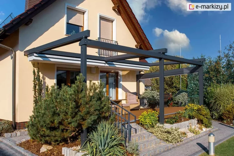 Nowoczesna pergola tarasowa przyścienna Solid o konstrukcji aluminiowej w kolorze antracytowym, przedstawiona ze zwiniętym, jasnym poszyciem tkaninowym, zamontowana przy beżowej elewacji domu jednorodzinnego. Aluminiowy stelaż opiera się na solidnych profilach pionowych, a zadaszenie segmentowe wsparte jest na poprzecznych belkach, tworząc zintegrowaną osłonę nad tarasem wykonanym z brązowych desek kompozytowych. Na taras prowadzą kamienne schody z szarego granitu wyposażone w metalową balustradę, a w strefie wypoczynkowej pod pergolą widoczne są plecione fotele ogrodowe z jasnymi poduszkami. Wokół tarasu znajduje się bogato zaaranżowany ogród z dużą sosną górską, juką oraz niskimi krzewami ozdobnymi posadzonymi na podłożu z kory sosnowej, oddzielonym od ścieżki z kostki brukowej kamiennymi krawężnikami. Wzdłuż traktu komunikacyjnego ustawiono nowoczesne lampy ogrodowe w formie srebrnych słupków, a w tle widoczne są okna budynku z opuszczonymi roletami zewnętrznymi oraz błękitne niebo z delikatnymi chmurami.