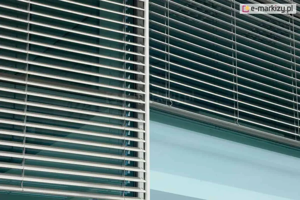 Zbliżenie na elewację nowoczesnego budynku biurowego z zamontowanymi zewnętrznymi żaluzjami fasadowymi Z90, o poziomych, aluminiowych listwach w kolorze srebrnym, które częściowo przesłaniają szklaną taflę okna. Żaluzje są ustawione pod kątem, co pozwala na kontrolowanie ilości wpadającego do wnętrza światła słonecznego, a pionowy profil prowadnicy listwowej oddziela dwa sąsiadujące ze sobą segmenty okienne, na których widoczne jest odbicie nieba i architektury w gładkiej powierzchni szyb, podkreślając industrialny i minimalistyczny charakter zastosowanego systemu zacieniania.