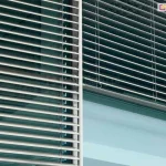 Zbliżenie na elewację nowoczesnego budynku biurowego z zamontowanymi zewnętrznymi żaluzjami fasadowymi Z90, o poziomych, aluminiowych listwach w kolorze srebrnym, które częściowo przesłaniają szklaną taflę okna. Żaluzje są ustawione pod kątem, co pozwala na kontrolowanie ilości wpadającego do wnętrza światła słonecznego, a pionowy profil prowadnicy listwowej oddziela dwa sąsiadujące ze sobą segmenty okienne, na których widoczne jest odbicie nieba i architektury w gładkiej powierzchni szyb, podkreślając industrialny i minimalistyczny charakter zastosowanego systemu zacieniania.