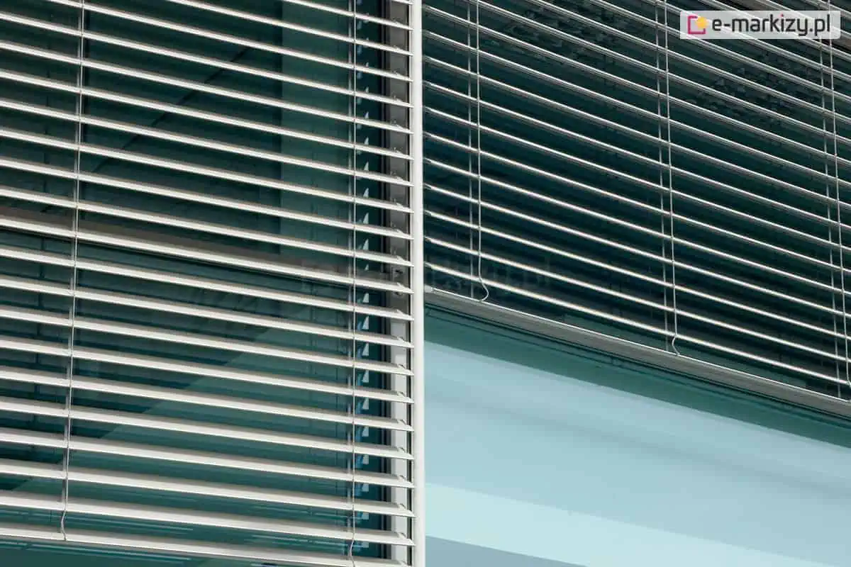 Zbliżenie na elewację nowoczesnego budynku biurowego z zamontowanymi zewnętrznymi żaluzjami fasadowymi Z90, o poziomych, aluminiowych listwach w kolorze srebrnym, które częściowo przesłaniają szklaną taflę okna. Żaluzje są ustawione pod kątem, co pozwala na kontrolowanie ilości wpadającego do wnętrza światła słonecznego, a pionowy profil prowadnicy listwowej oddziela dwa sąsiadujące ze sobą segmenty okienne, na których widoczne jest odbicie nieba i architektury w gładkiej powierzchni szyb, podkreślając industrialny i minimalistyczny charakter zastosowanego systemu zacieniania.