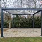 Nowoczesna pergola tarasowa wolnostojąca SB350 Selt o konstrukcji aluminiowej w kolorze ciemnoszarym (antracytowym), zamontowana na podłożu z jasnych płyt betonowych. Zadaszenie pergoli stanowią ruchome lamele w kolorze białym, ustawione pod kątem, co pozwala na regulację dopływu światła słonecznego. Konstrukcja opiera się na czterech solidnych, kwadratowych słupach przytwierdzonych do podłoża za pomocą stóp montażowych. W tle widoczny jest ogród z gęstą roślinnością, obejmującą iglaki, krzewy oraz wysokie, bezlistne drzewa liściaste w okresie wczesnowiosennym lub jesiennym. Po prawej stronie zdjęcia znajduje się duża donica z rośliną, a po lewej młode drzewka zabezpieczone tyczkami bambusowymi.