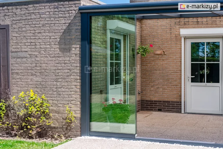 Zdjęcie przedstawia nowoczesną zabudowę ganku lub tarasu z wykorzystaniem przesuwnych drzwi szklanych Auris, zamontowanych w grafitowej, aluminiowej ramie konstrukcyjnej. Bezramowe panele ze szkła hartowanego tworzą przezroczystą ścianę osłonową, która jest częściowo zsunięta, ukazując wejście na wykafelkowany taras. W tle widoczna jest elewacja budynku wykonana z tradycyjnej, jasnobrązowej cegły klinkierowej, w której osadzono białe drzwi wejściowe z przeszkleniami i szprosami, wyposażone w zewnętrzną roletę zabezpieczającą. Wewnątrz zadaszonej przestrzeni, przy ścianie, stoi donica z kwitnącymi na czerwono pelargoniami, a wyżej zawieszona jest mniejsza kaskadowa roślina doniczkowa oraz ozdobna tabliczka. Po lewej stronie kadru znajduje się fragment prostopadłej ściany z szarej cegły, u podnóża której rośnie zielony krzew z żółtymi akcentami liści. Przed szklaną zabudową przebiega opaska z jasnego żwiru oraz fragment zielonego trawnika, co podkreśla integrację nowoczesnej konstrukcji ogrodowej z otoczeniem domu.