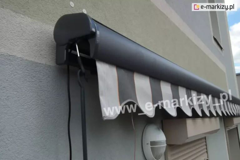 Markiza Palladio w kolorze konstrukcji srebrzysty grafit z widocznym po lewej stronie sterowaniem awaryjnym za pomocą korby oraz przewodem zasilania napędu elektrycznego. Markiza zawieszona na ścianie o dwukolorowej elewacji niebiesko-zielonej. Falowana falbana w pasy w dwóch odcieniach zieleni z delikatnym czerwonym paskiem pomiędzy nimi sięga do krawędzi lampy oraz skrzynek rolet zewnętrznych zamocowanych poniżej na oknach.