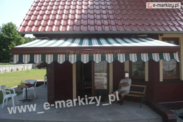 Zdjęcie prezentuje efekt końcowy montażu markizy tarasowej na budynku o ciemnoczerwonej elewacji, pokrytym dachówką ceramiczną w kolorze ceglastym. Markiza Palladio Aluprof jest w pełni rozwinięta, a jej poszycie wykonane jest z materiału w pionowe pasy w kolorach białym, ciemnozielonym oraz szarym. Dolna krawędź tkaniny wykończona jest dekoracyjną, falistą falbaną, która powtarza kolorystykę i wzór głównej połaci. Konstrukcja markizy, w tym belka frontowa oraz kaseta, wykonana jest w kolorze ciemnobrązowym, co współgra z barwą elewacji oraz stolarki okiennej. Pod zadaszeniem, na betonowym tarasie, znajduje się strefa wypoczynkowa z drewnianą ławką, na której siedzi mężczyzna. Po lewej stronie tarasu ustawione są białe, plastikowe krzesła ogrodowe oraz mały stolik. W tle widoczny jest zadbany trawnik, niskie ogrodzenie posesji oraz ściana zielonych drzew pod jasnym niebem. Markiza skutecznie zacienia wejście tarasowe i okna parteru, tworząc komfortową przestrzeń do odpoczynku na zewnątrz.