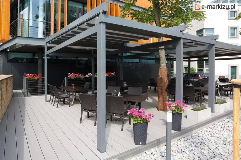 Nowoczesna, wielomodułowa pergola tarasowa Solid Aluprof o konstrukcji aluminiowej w kolorze antracytowym, zainstalowana jako ogródek letni przy nowoczesnym budynku. Zadaszenie wykonane jest z jasnoszarej tkaniny rzymskiej, która jest rozsunięta na systemie prowadnic. Pod pergolą, na tarasie z jasnych desek kompozytowych, ustawiono liczne czarne stoliki restauracyjne z dopasowanymi fotelami z technorattanu. Konstrukcja została zaprojektowana z uwzględnieniem rosnącego wewnątrz drzewa, którego pień zabezpieczono brązową osłoną. Przestrzeń jest udekorowana wysokimi, czarnymi donicami z różowymi kwiatami oraz białymi, podłużnymi skrzyniami z roślinnością. W tle widoczny jest fragment budynku z elewacją z drewna i szkła oraz pas jasnego żwiru z obrzeżem. Zdjęcie wykonane w pełnym świetle dziennym.