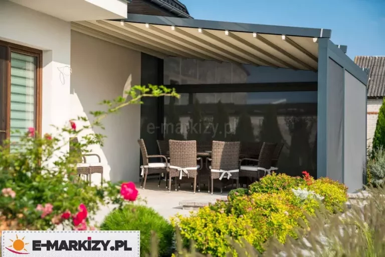 Widok na jadalnię tarasową urządzoną pod nowoczesną pergolą o ciemnoantracytowej konstrukcji z jasnym, segmentowym dachem tkaninowym, w który wkomponowano punktowe oświetlenie LED rozmieszczone symetrycznie na profilach poprzecznych. Pod zadaszeniem znajduje się zestaw mebli ogrodowych składający się z dużego stołu oraz ośmiu krzeseł wykonanych z brązowego technorattanu z jasnymi poduszkami na siedziskach. Tył oraz bok pergoli zostały przesłonięte pionowymi roletami zewnętrznymi typu screen w ciemnym kolorze, które zapewniają prywatność i ochronę przed słońcem, jednocześnie zachowując częściową przezierność. Całość kompozycji dopełnia otoczenie bujnej roślinności ogrodowej z kwitnącymi różami na pierwszym planie oraz starannie przystrzyżonymi krzewami ozdobnymi wzdłuż krawędzi jasnego, kamiennego tarasu.