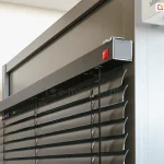 Zbliżenie na górny profil oraz mechanizm montażowy żaluzji poziomej C50 Slim Aluprof o nowoczesnym designie. Zdjęcie prezentuje czarne, aluminiowe lamele o wygiętym profilu w kształcie litery C, zawieszone na precyzyjnym systemie drabinek sznurkowych. Górna rynna żaluzji jest osłonięta elegancką, szeroką maskownicą w kolorze antracytowym o matowym wykończeniu, która jest stabilnie zamontowana do ramy okiennej. Na bocznej krawędzi maskownicy widoczny jest subtelny, czerwony logotyp producenta oraz metalowe elementy wykończeniowe. Detal podkreśla wysoką jakość spasowania komponentów oraz techniczny charakter systemu osłonowego, idealnie komponującego się z nowoczesną stolarką otworową. Tło stanowi neutralna, jasna ściana oraz fragment profilu aluminiowego, co pozwala w pełni skupić uwagę na konstrukcji samej żaluzji.