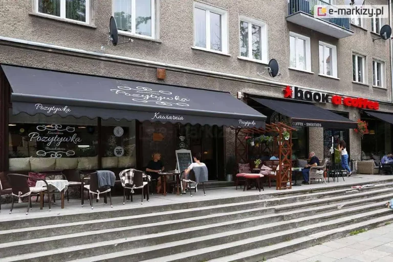 Czarna markiza reklamowa z ozdobnymi białymi napisami, osłaniająca ogródek kawiarniany w parterze miejskiej kamienicy. Na głównym poszyciu oraz na pofalowanej falbanie widnieje estetyczny nadruk logo kawiarni "Pozytywka" wykonany ozdobną czcionką z dekoracyjnymi zawijasami. Obok znajduje się kolejna, mniejsza osłona pod szyldem "book a coffee". Pod markizami ustawiono liczne stoliki, wiklinowe i metalowe krzesła z poduszkami oraz koce dla gości. Witryny lokalu są przeszklone, a na jednej z nich powtórzono logotyp kawiarni. Przed wejściem do lokali znajdują się szerokie, betonowe schody prowadzące na poziom chodnika. W ogródku widoczni są klienci siedzący przy stolikach oraz drewniane konstrukcje pergoli z donicami kwiatowymi. Elewacja budynku jest szara, o chropowatej strukturze, z widocznymi powyżej oknami mieszkalnymi i antenami satelitarnymi. Całość tworzy przytulną przestrzeń komercyjną zintegrowaną z miejską architekturą.