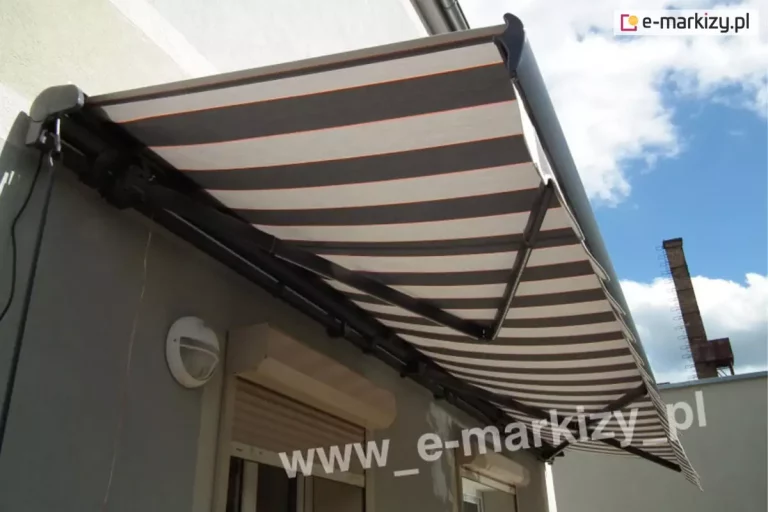 Ujęcie od boku ukazujące nowoczesną markizę tarasową Palladio Aluprof, w wysuwie, zamontowaną na elewacji budynku mieszkalnego. Poszycie markizy charakteryzuje się wzorem w szerokie, poziome pasy w kolorze szarym i białym, które są oddzielone od siebie cienkimi, pomarańczowymi liniami akcentowymi. Materiał jest napięty na solidnej, ciemnej konstrukcji mechanicznej, wykonanej z aluminium lakierowanego proszkowo na kolor ciemny grafit. Pod spodem widoczne są przegubowe ramiona składane, które wspierają tkaninę i odpowiadają za jej stabilność po rozłożeniu. Mechanizm mocujący markizę jest przytwierdzony do szarej ściany budynku tuż nad oknem wyposażonym w zewnętrzną roletę okienną w kolorze jasnobeżowym. Obok okna, na elewacji, znajduje się biała, okrągła lampa zewnętrzna o półkolistym kloszu. W tle widać fragment błękitnego nieba z rozproszonymi białymi chmurami oraz zarys wysokiego, ceglanego komina przemysłowego w oddali, co sugeruje lokalizację obiektu w terenie zurbanizowanym. Całość kompozycji ukazuje detale techniczne montażu oraz sposób funkcjonowania ochrony przeciwsłonecznej w warunkach dziennych.