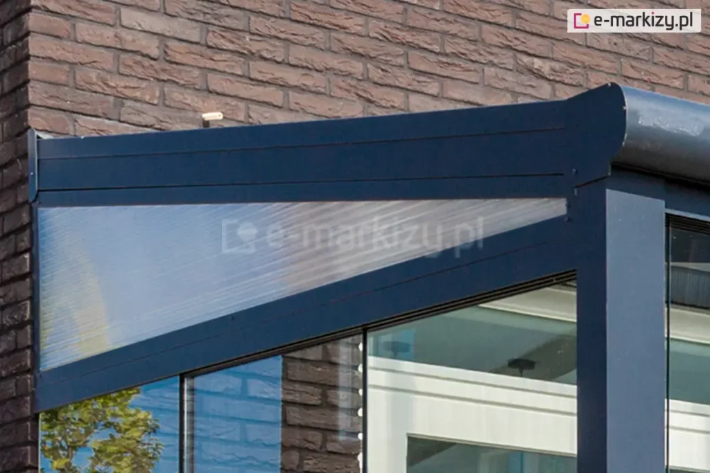 Prezentacja techniczna bocznego klina wypełniającego, zaprojektowanego specjalnie dla systemów zadaszeń Lunaro, wykonanego w nowoczesnym wykończeniu antracytowym. Element ten pełni funkcję stabilnego trójkątnego domknięcia przestrzeni między skośnym profilem dachowym a elewacją budynku, która w tym przypadku została wykonana z surowej, brązowej cegły klinkierowej. Aluminiowa rama klina, zabezpieczona lakierem proszkowym o matowej fakturze, gwarantuje wysoką trwałość konstrukcyjną oraz pełną odporność na zmienne warunki atmosferyczne, idealnie komponując się z ciemną kolorystyką pionowych słupów podporowych. Wewnątrz ramy umieszczono panel z poliwęglanu wielokomorowego, który dzięki swojej charakterystycznej, pionowo żebrowanej strukturze zapewnia doskonałe doświetlenie wnętrza tarasu, jednocześnie zachowując wysokie parametry wytrzymałościowe i sztywność całego elementu. Pod dolną krawędzią klina widoczne są elementy systemu szyb przesuwnych, natomiast w głębi kadru można dostrzec białe profile sufitowe z zainstalowanym punktowym oświetleniem, co podkreśla kompleksowy charakter zabudowy strefy wypoczynkowej przy jednoczesnym zapewnieniu doskonałej szczelności konstrukcji przed wiatrem i deszczem.