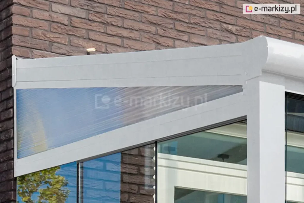 Zbliżenie z zewnątrz na narożny klin wykończeniowy z białą ramą, zamontowany w systemie zadaszenia tarasowego Lunaro. Element ten ma formę trójkąta prostokątnego i stanowi niezbędny moduł domykający otwartą przestrzeń pomiędzy nachylonym profilem dachu a pionową powierzchnią ściany budynku, wykończonej ciemną cegłą klinkierową. Rama klina, polakierowana proszkowo na kolor biały, wykazuje wysoką odporność na działanie czynników atmosferycznych, takich jak promieniowanie UV czy wilgoć. Wypełnienie stanowi arkusz wielokomorowego poliwęglanu o przejrzystej strukturze, który charakteryzuje się subtelnym, jasnoniebieskim zabarwieniem oraz podłużnym ryflowaniem komór, poprawiającym parametry wytrzymałościowe materiału. Pod klinem widoczne jest zintegrowane przeszklenie przesuwne tarasu oraz fragment białych profili sufitowych. Całość montażu prezentuje estetyczne połączenie nowoczesnej konstrukcji zadaszenia z elewacją, zapewniając skuteczną ochronę przed wiatrem i deszczem przy zachowaniu pełnej spójności wizualnej z jasną kolorystyką słupów wsporczych i ramy dachowej.