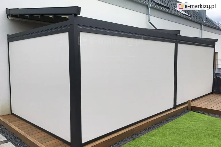 Zdjęcie przedstawia model pergoli tarasowej Square przyściennej o konstrukcji aluminiowej w kolorze antracytowym, która została niemal całkowicie zabudowana pionowymi przesłonami bocznymi. Pergola jest zamontowana do białej elewacji budynku mieszkalnego i posadowiona na tarasie wykonanym z naturalnych, brązowych desek drewnianych, które są otoczone pasem ciemnego żwiru oraz zadbanym, zielonym trawnikiem. Głównym elementem widocznym na zdjęciu są trzy duże, prostokątne ekrany boczne (refleksol Zip l Zip U) w kolorze jasnokremowym lub bardzo jasnym szarym, które są w pełni rozwinięte. Przesłony te są szczelnie osadzone w prowadnicach bocznych zintegrowanych ze słupami pergoli, tworząc jednolitą i gładką ścianę chroniącą wnętrze przed wiatrem, słońcem oraz wzrokiem osób trzecich. Konstrukcja dachu pergoli jest lekko nachylona, a nad przesłonami bocznymi widoczna jest kaseta systemu oraz fragmenty poprzecznych belek podtrzymujących zadaszenie. W tle, po prawej stronie, widać fragment sąsiedniego budynku z dachem pokrytym ciemną dachówką oraz rynnę odprowadzającą wodę z elewacji głównego domu. Całość kompozycji podkreśla minimalistyczny i funkcjonalny charakter zabudowy tarasu, zapewniający pełną prywatność użytkownikom.