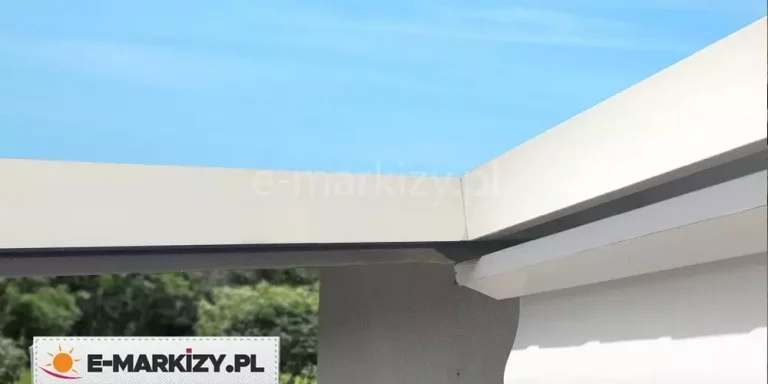 Detaliczne zbliżenie na narożne połączenie dwóch białych belek konstrukcyjnych aluminiowej pergoli tarasowej Luxo na tle bezchmurnego, błękitnego nieba. Zdjęcie ukazuje precyzyjne, prostopadłe styki poziomych profili, pod którymi widoczna jest ciemna uszczelka lub element maskujący system odprowadzania wody. Pod główną ramą zamontowano mniejszy, biały profil wykończeniowy o lekko zaokrąglonym kształcie, osłaniający krawędź zwiniętego poszycia dachowego. Konstrukcja charakteryzuje się gładką, matową powierzchnią lakierowaną proszkowo. W dolnej części kadru, poza ostrością, widoczny jest zarys zielonych koron drzew oraz szary fragment elewacji budynku, co podkreśla wysokość montażu i minimalistyczny design systemu.