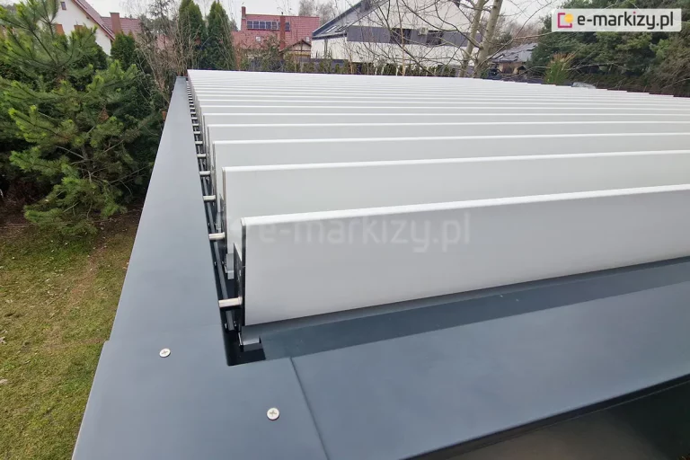 Widok z góry na krawędź dachu pergoli bioklimatycznej Sb350 Aluprof prezentujący system ruchomych lameli aluminiowych w kolorze nialym. Lamele są ustawione w pozycji lekko uchylonej, co odsłania ich mechanizm obrotowy oraz stalowe trzpienie mocujące je do antracytowej ramy konstrukcyjnej. Na górnej powierzchni szerokiego profilu bocznego widoczne są srebrne śruby montażowe. W tle, pod kątem, rozpościera się widok na ogród z zielonymi iglakami, trawnikiem oraz fragmentami sąsiedniej zabudowy jednorodzinnej z białymi elewacjami i ciemnymi dachami pod zachmurzonym niebem.