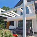 Pergola drewniana Moza z dachem rzymskim Supro zwiniętym na jedną stronę, dzięki czemu krokwie dachowe zostały odsłonięte i widoczny jest system sterowania ręcznego sznurkowego. pod dachem ustawiono meble tarasowe z szarymi poduchami. Na pierwszym tle kolorowe rośliny ogrodowe, a nad pergolą niebieskie bezchmurne niebo.