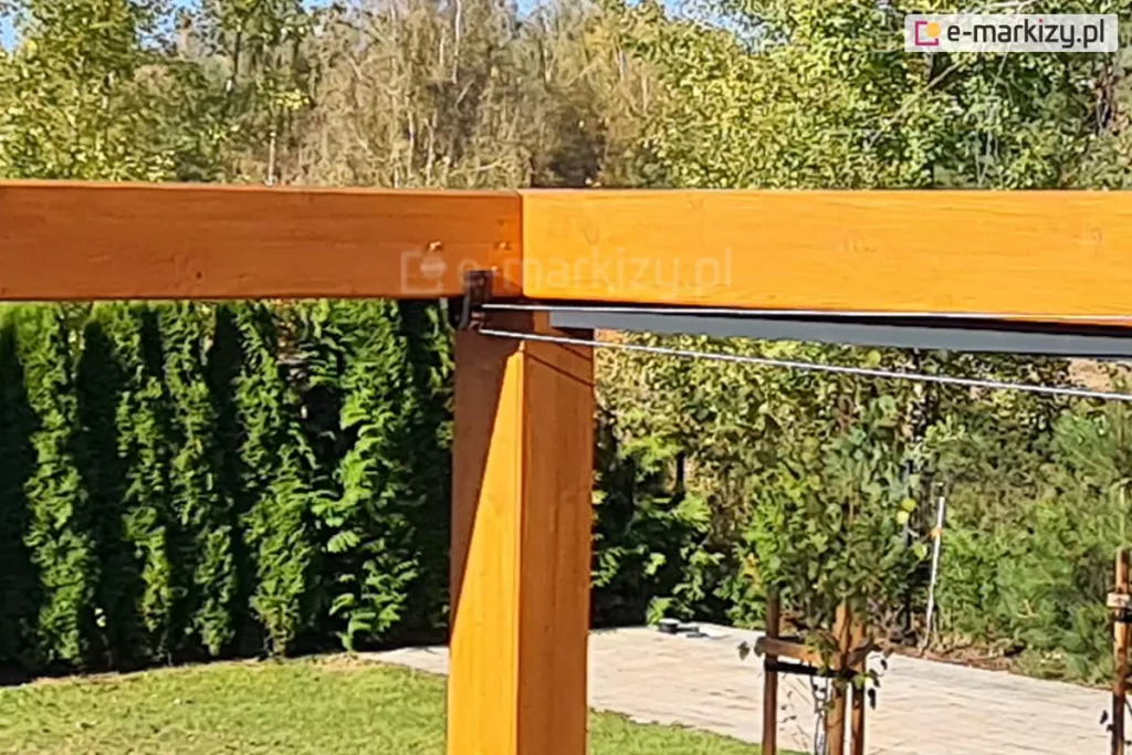 Konstrukcja drewniana nad taras koro z rozwijanym zadaszeniem tkaninowym supro elektrycznym, którego elementy przesuwane są za pomocą linek stalowych. W tle bogata, zielona roślinność ogrodu oraz wczesnowiosenne drzewa pozbawione jeszcze liści.