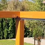 Konstrukcja drewniana nad taras koro z rozwijanym zadaszeniem tkaninowym supro elektrycznym, którego elementy przesuwane są za pomocą linek stalowych. W tle bogata, zielona roślinność ogrodu oraz wczesnowiosenne drzewa pozbawione jeszcze liści.