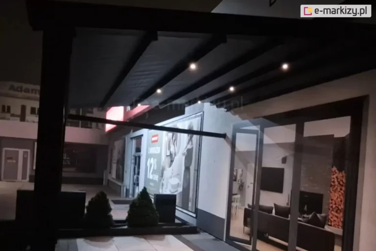 Zdjęcie prezentuje pergolę tarasową Square zamontowaną do elewacji budynku, sfotografowaną w warunkach nocnych. Konstrukcja pergoli jest wykonana z grubych profili aluminiowych w kolorze czarnym lub bardzo ciemnym antracycie. Zadaszenie składa się z rozsuwanego, ciemnego materiału, który jest rozwinięty i ułożony w równoległe segmenty. Pod poszyciem, na poprzecznych belkach konstrukcyjnych, zamontowano system zintegrowanego oświetlenia LED w postaci jasnych, punktowych diod emitujących ciepłe światło, które oświetlają przestrzeń pod pergolą. Pergola jest przytwierdzona do ściany budynku, w której znajdują się duże, przeszklone drzwi tarasowe oraz witryna. Przez szyby widoczne jest nowocześnie urządzone wnętrze salonu z ciemną sofą, telewizorem zamontowanym na ścianie oraz dekoracyjnym stosem drewna kominkowego ułożonym we wnęce. Na zewnątrz, bezpośrednio pod zadaszeniem, znajdują się niskie donice z małymi, przyciętymi krzewami iglastymi w kształcie stożków. W tle, po lewej stronie, widoczny jest fragment innego budynku z podświetlonym czerwonym szyldem reklamowym oraz napisem, co sugeruje lokalizację w przestrzeni komercyjnej lub ekspozycyjnej. Całość kompozycji podkreśla kontrast między ciemną bryłą konstrukcji a punktami świetlnymi oraz rozświetlonym wnętrzem domu.