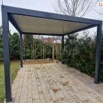 Wolnostojąca pergola ogrodowa SB350 Selt o nowoczesnej konstrukcji aluminiowej w kolorze antracytowym, zainstalowana na podłożu z jasnej kostki brukowej. Dach pergoli stanowią zamknięte lamele bioklimatyczne w kolorze białym, tworzące pełne, szczelne zadaszenie. Konstrukcja opiera się na czterech solidnych, kwadratowych słupach z widocznymi stopami montażowymi przytwierdzonymi do podłoża. W tle znajduje się gęsta roślinność ogrodowa: wysokie krzewy iglaste, sosna oraz młode brzozy zabezpieczone drewnianymi palikami. Po lewej stronie widoczny jest fragment zielonego trawnika, a po prawej w oddali zabudowania mieszkalne pod pochmurnym niebem. Na lewym słupie narożnym widoczna jest pionowa czarna antena sterowania radiowego.