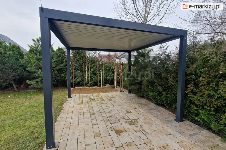 Wolnostojąca pergola ogrodowa SB350 Selt o nowoczesnej konstrukcji aluminiowej w kolorze antracytowym, zainstalowana na podłożu z jasnej kostki brukowej. Dach pergoli stanowią zamknięte lamele bioklimatyczne w kolorze białym, tworzące pełne, szczelne zadaszenie. Konstrukcja opiera się na czterech solidnych, kwadratowych słupach z widocznymi stopami montażowymi przytwierdzonymi do podłoża. W tle znajduje się gęsta roślinność ogrodowa: wysokie krzewy iglaste, sosna oraz młode brzozy zabezpieczone drewnianymi palikami. Po lewej stronie widoczny jest fragment zielonego trawnika, a po prawej w oddali zabudowania mieszkalne pod pochmurnym niebem. Na lewym słupie narożnym widoczna jest pionowa czarna antena sterowania radiowego.