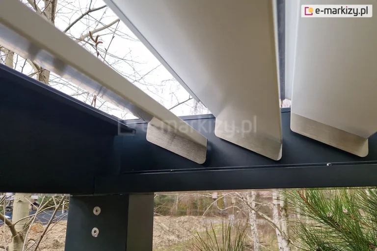 Techniczne zbliżenie na wewnętrzny mechanizm wspornikowy dachu pergoli bioklimatycznej Sb350 Aluprof. Zdjęcie przedstawia aluminiowe lamele w kolorze białym, które są wyposażone w metalowe, srebrzyste zakończenia o szczotkowanej strukturze. Lamele są ustawione w pozycji uchylonej, ukazując sposób ich osadzenia w bocznej belce konstrukcyjnej o barwie antracytowej, pod którą znajduje się rynna odpływowa. Na pionowym słupie nośnym widoczne są dwa srebrne gniazda śrub montażowych. W tle, pod kątem, widoczne są gałęzie drzew iglastych oraz fragmenty bezlistnych brzóz, co podkreśla ogrodowy charakter instalacji. Fotografia skupia się na precyzji spasowania elementów ruchomych z ramą stałą pergoli.