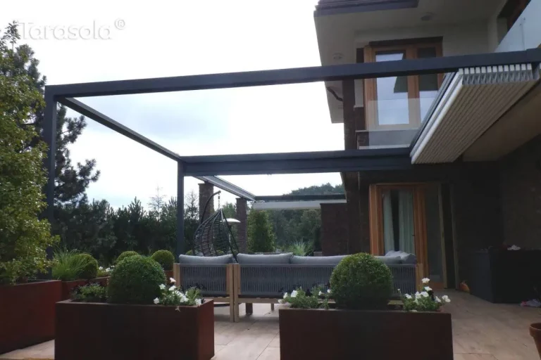 Zdjęcie przedstawia widok z boku na przyścienną pergolę tarasową Tarasola Flat o konstrukcji aluminiowej w kolorze antracytowym, której dach tkaninowy w kolorze jasnokremowym jest niemal całkowicie zwinięty w stronę elewacji budynku. Tkanina dachu spoczywa w formie ciasno złożonego pakietu pod dedykowanym daszkiem ochronnym, co odsłania pełną wysokość ramy i konstrukcję dachu płaskiego. Pergola przylega do elewacji budynku wykonanej z ciemnej cegły klinkierowej, z widocznym narożnym przeszkleniem i jasnym tynkiem na wyższej kondygnacji. Pod zadaszeniem znajduje się strefa wypoczynkowa z nowoczesną sofą ogrodową o szarej plecionej strukturze, ustawioną na tarasie wyłożonym jasnymi płytkami. Aranżację uzupełniają duże, podłużne donice ze stali corten o rdzawym odcieniu, w których rosną formowane w kule krzewy oraz białe kwiaty. W tle widoczny jest gęsty ogród z wysokimi drzewami iglastymi i liściastymi pod zachmurzonym niebem.